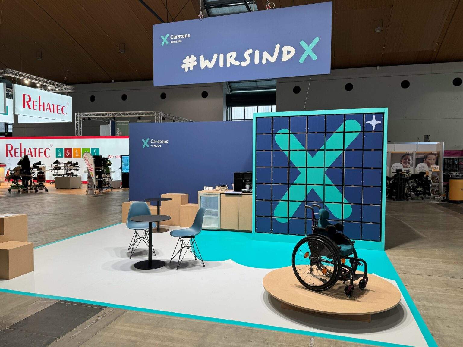 XCarstens trade fair stand with a “#WIRSINDX” banner and a large X wall, with a wheelchair on a platform in front